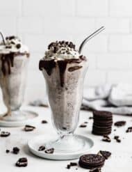 Two tall glasses full of Oreo milkshake topped with whipped cream and crushed Oreos.
