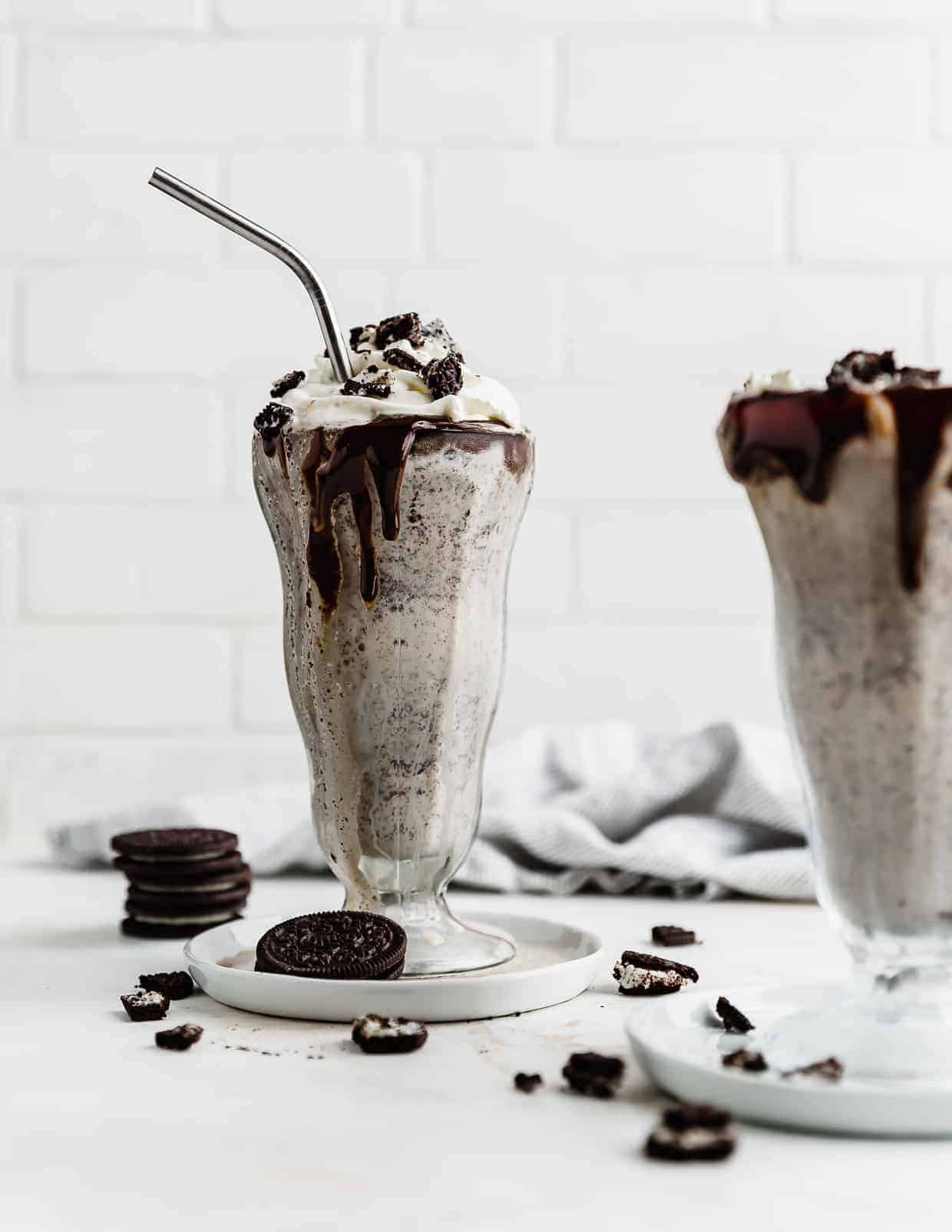 An oreo milkshake in a large glass cup, against a white brick background. 