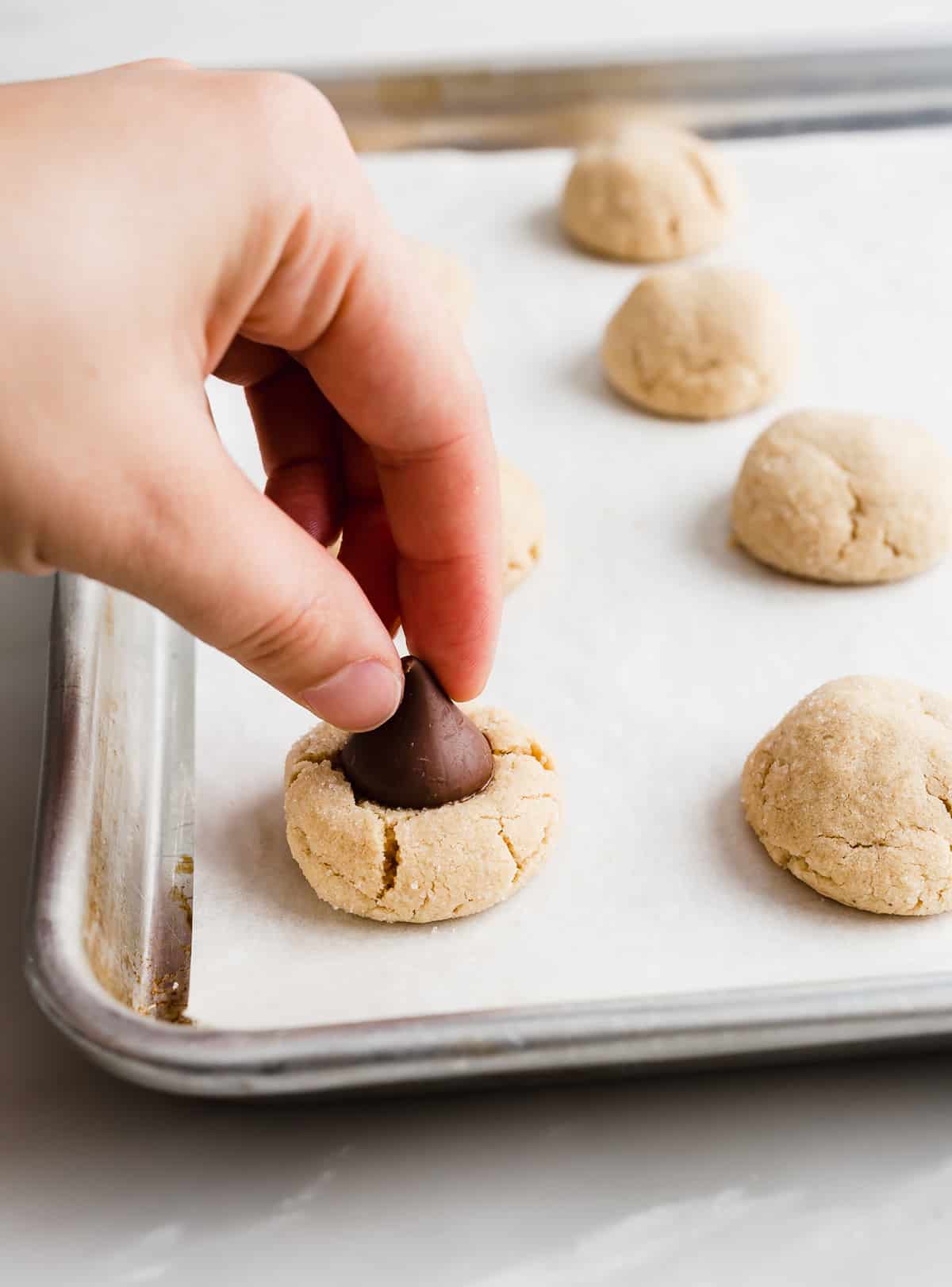 A hand pressing a chocolate Hershey's kiss into a peanut butter cookie on a baking sheet.