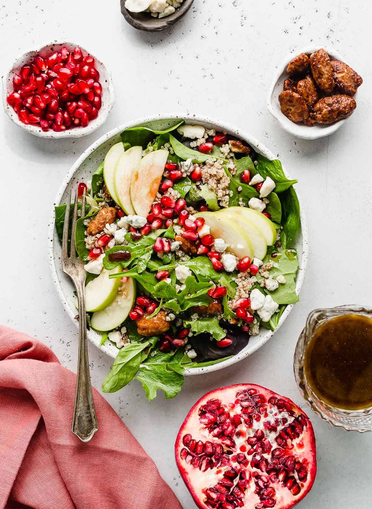 Pomegranate Salad on a white background with pomegranate dressing, nuts, red napkin, and fresh pomegranate surrounding the salad.