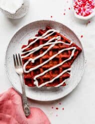 A white plate with 2 red velvet waffles on it drizzled in a white cream cheese glaze, against a white background.