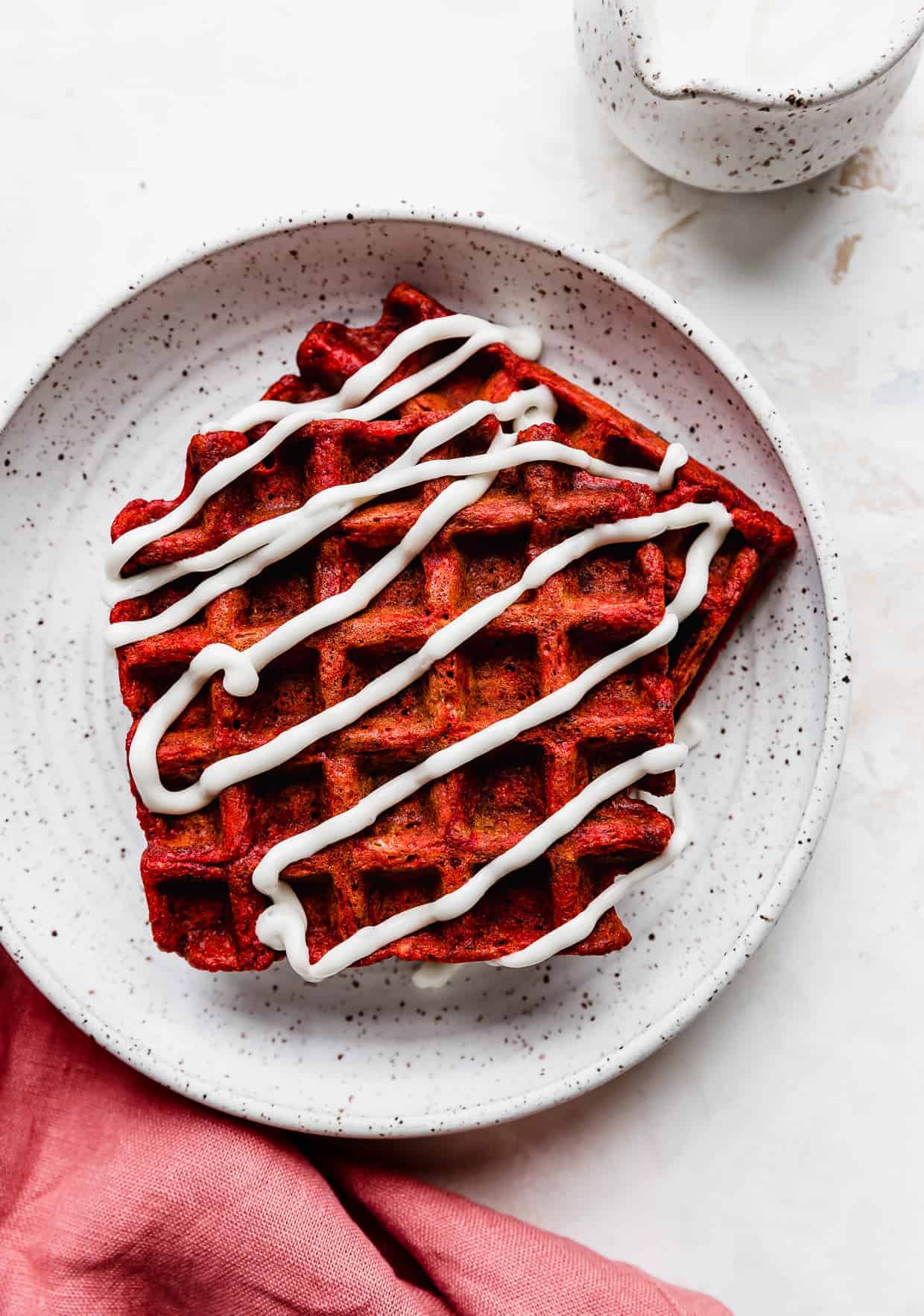 Close up of square red velvet waffles on a white plate with cream cheese glaze overtop of the waffles. 