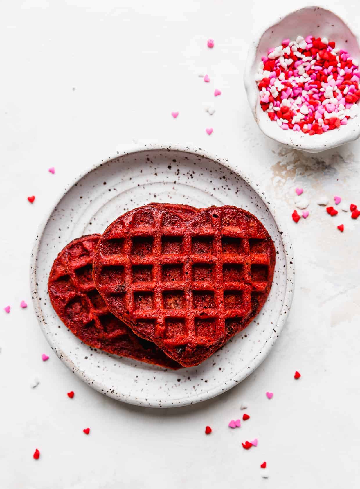 A heart shaped red velvet waffle on a white plate against a white background.