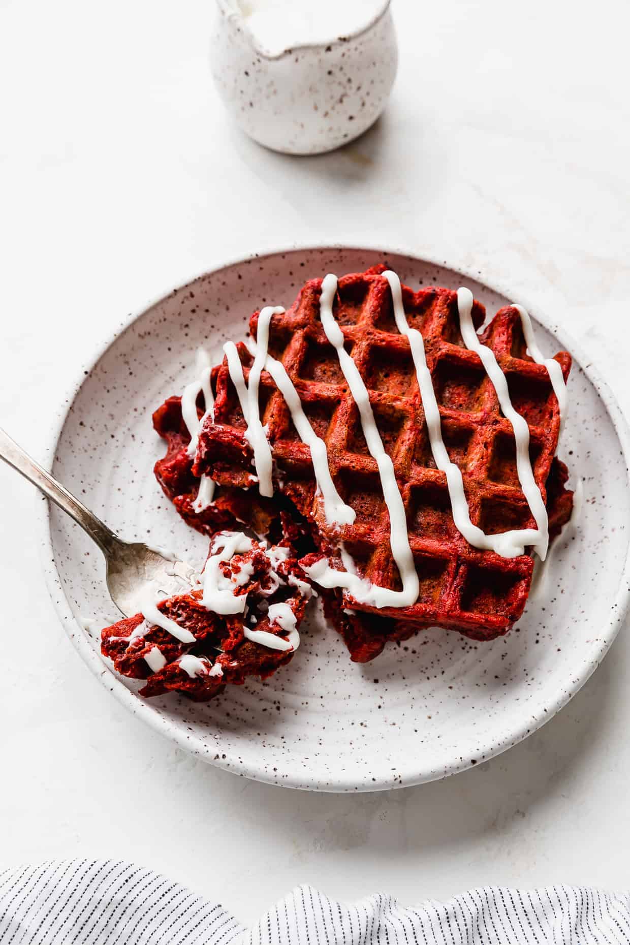 Two red velvet waffles drizzled with a white glaze on a white plate, against a white background.