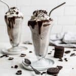 Two milkshake glasses filled with the best Oreo Milkshake recipe, against a white brick background with Oreo crumbs scattered around the cups.