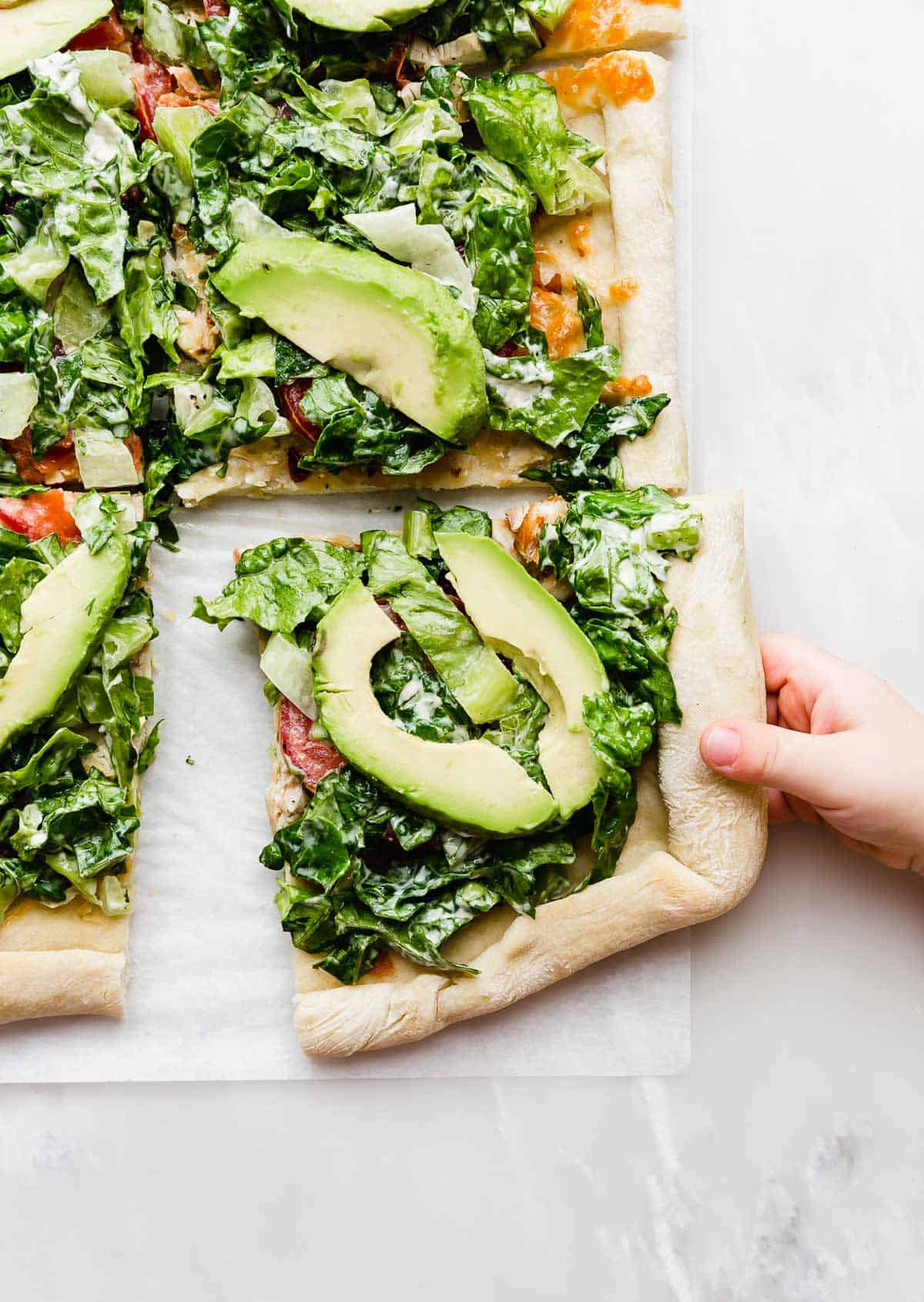 A hand grabbing a square slice of CPK Club Pizza that's topped with fresh avocado slices and lettuce.