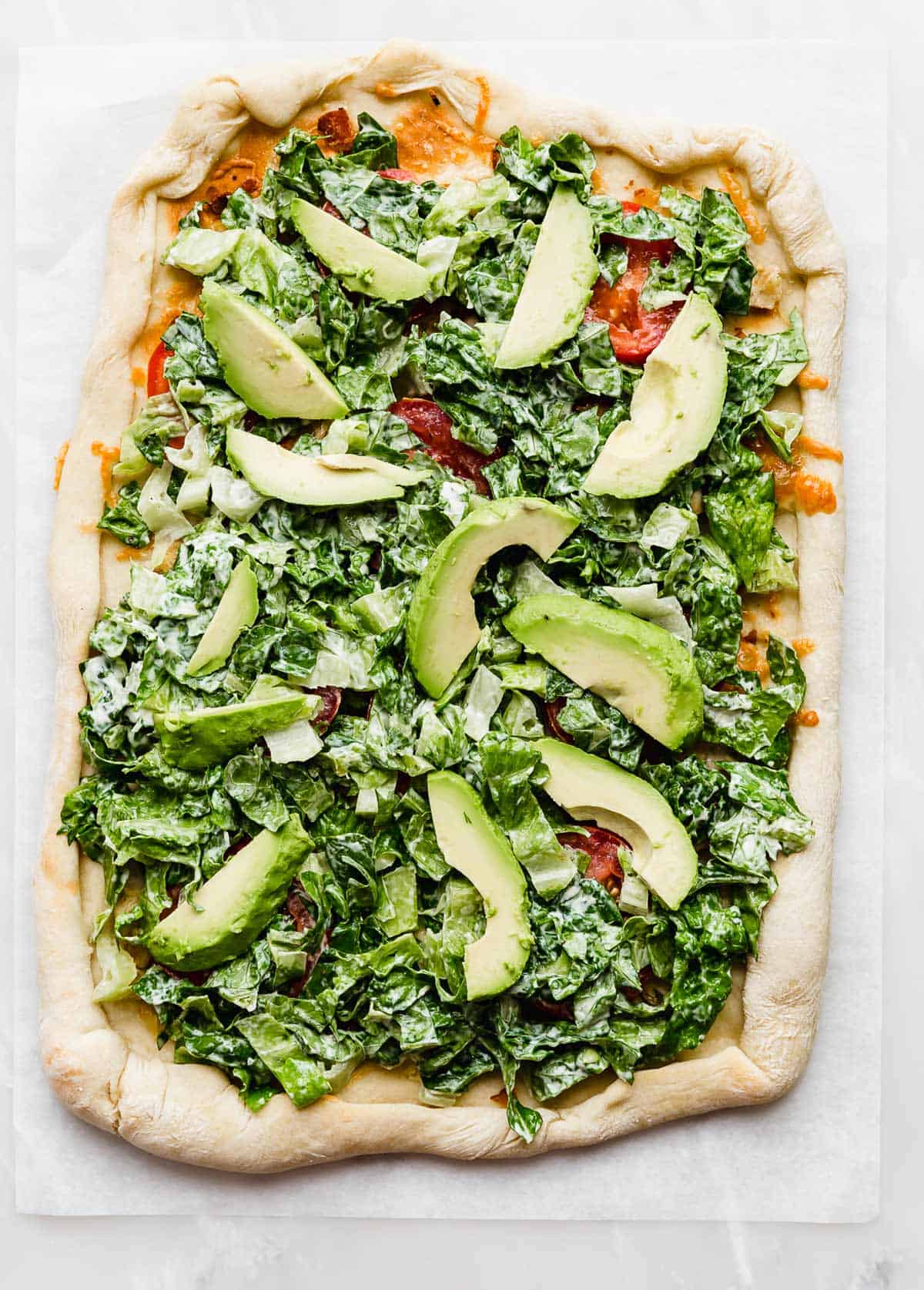 A rectangle type pizza topped with lettuce and avocado slices on a white background.