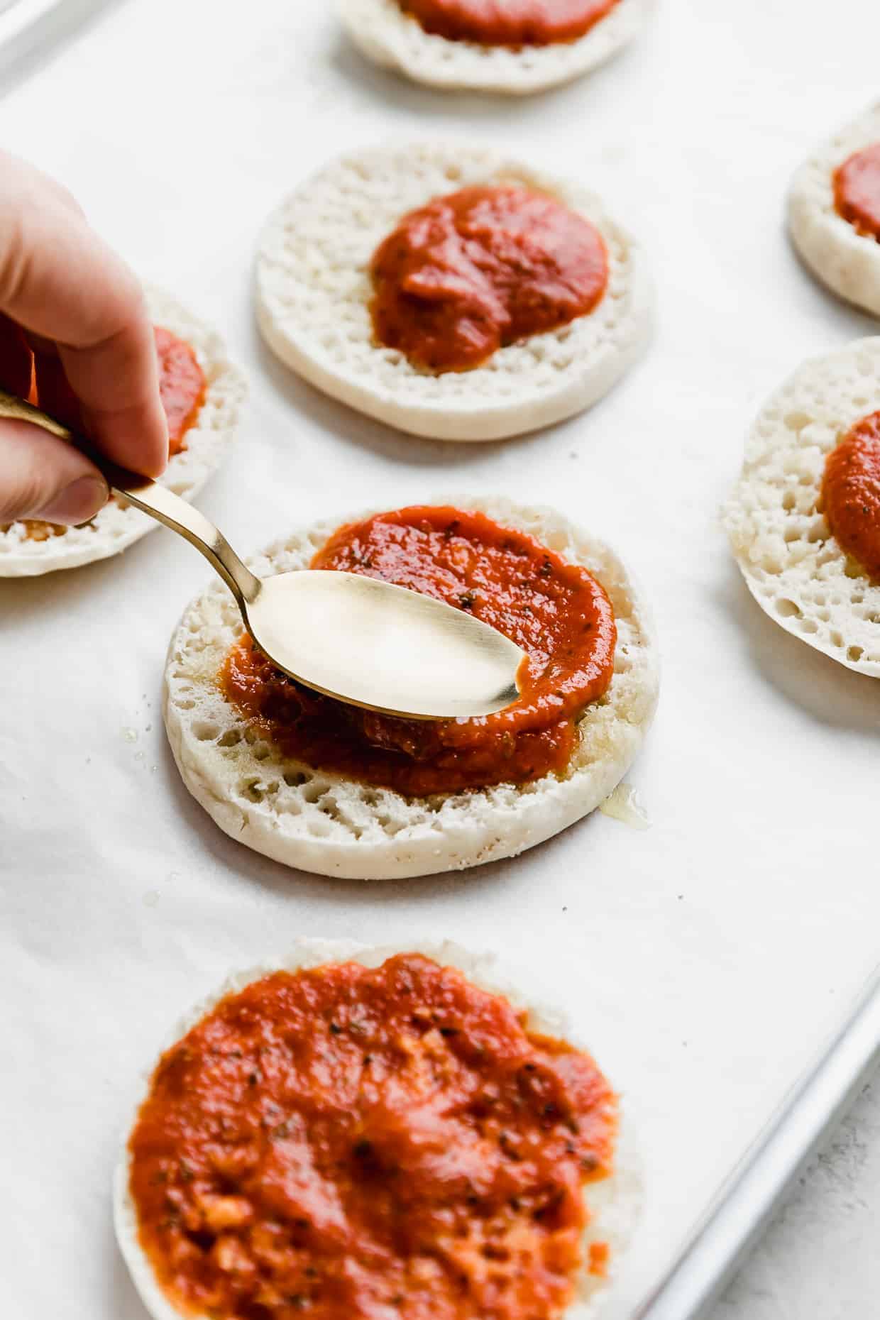A spoon spreading pizza sauce on a half of an English muffin. 