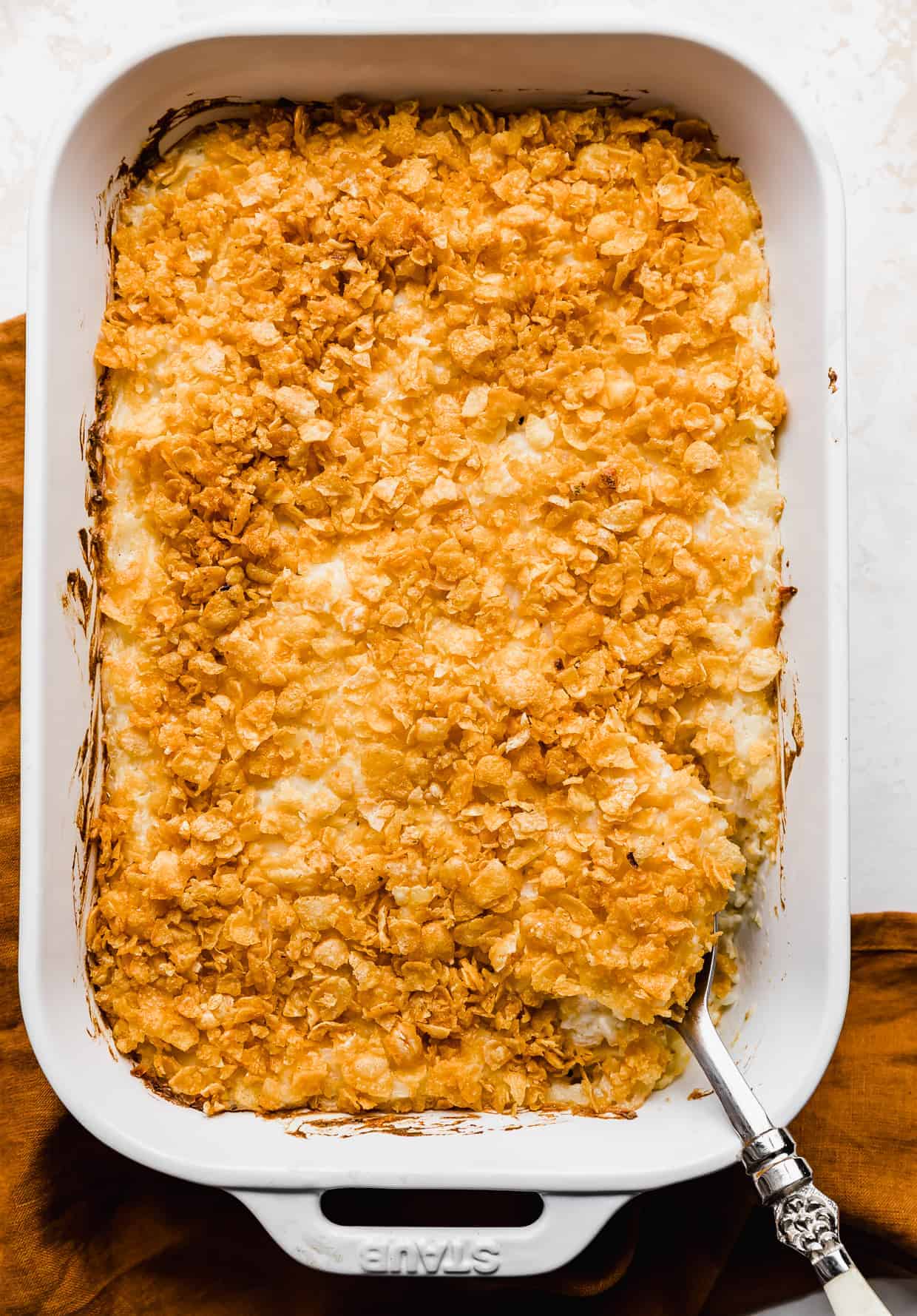Baked funeral potatoes topped with golden orange cornflakes, with a serving spoon scooping up a helping of potatoes. 