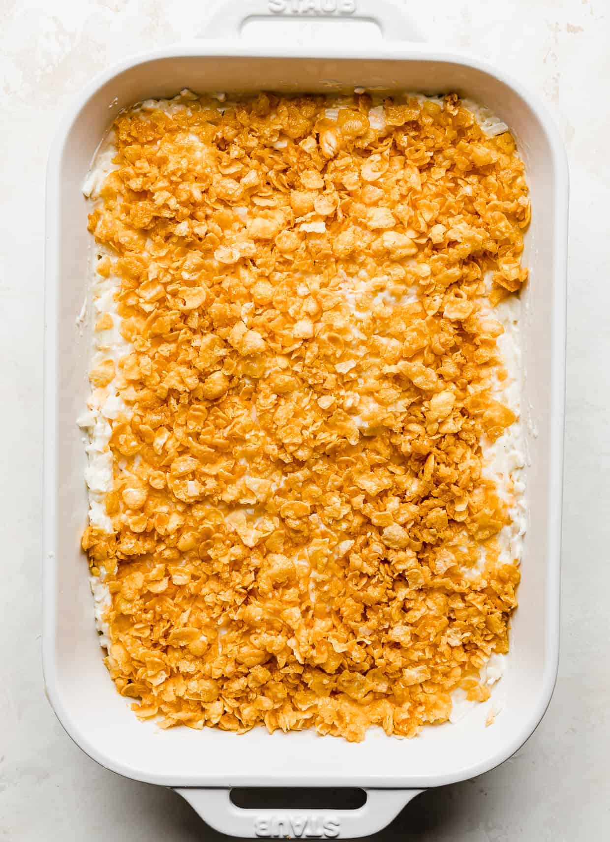 A casserole dish full of unbaked funeral potatoes topped with crushed cornflakes, against a white background.