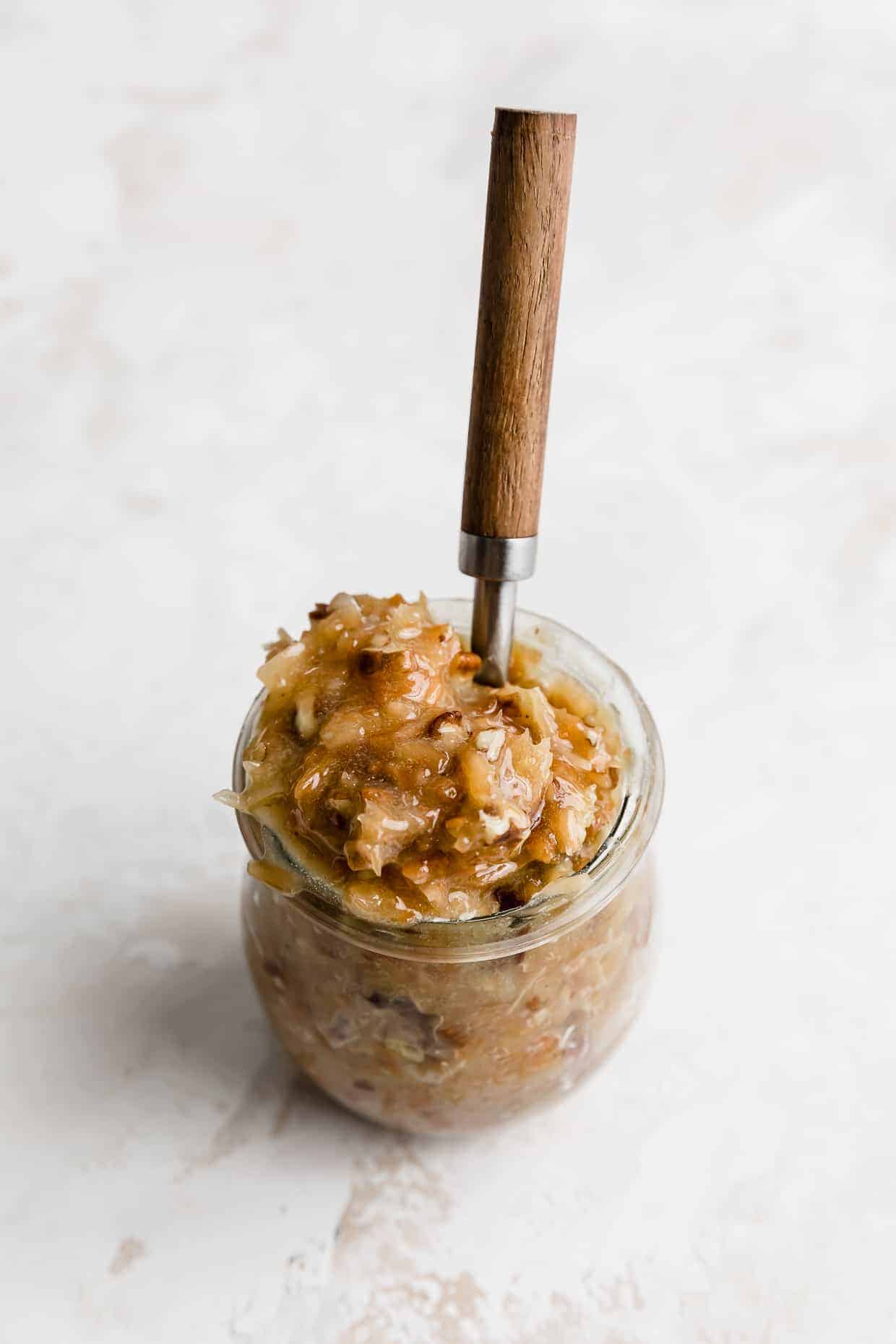 A small glass jar full of German Chocolate Cake frosting with a spoon sticking in the jar too.