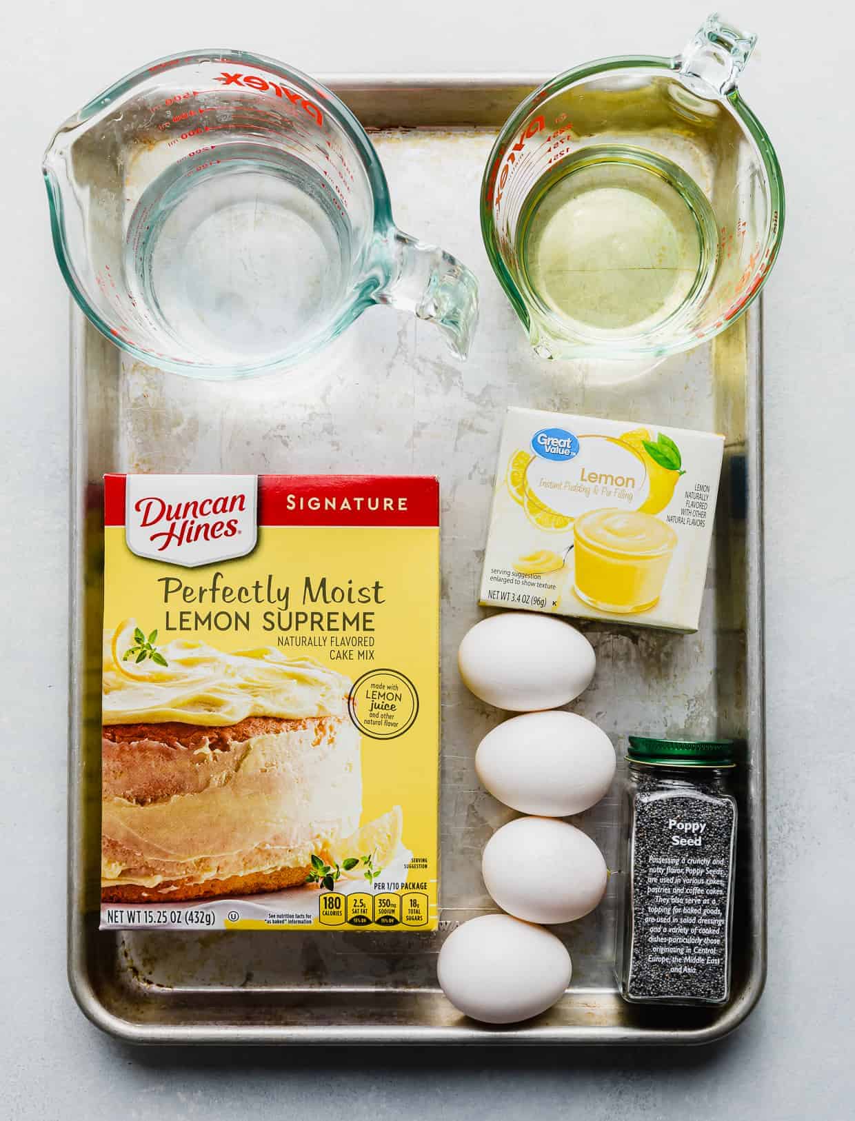Ingredients used to make lemon bread on a baking sheet: lemon cake mix, water, oil, eggs, lemon pudding mix, and poppy seeds.