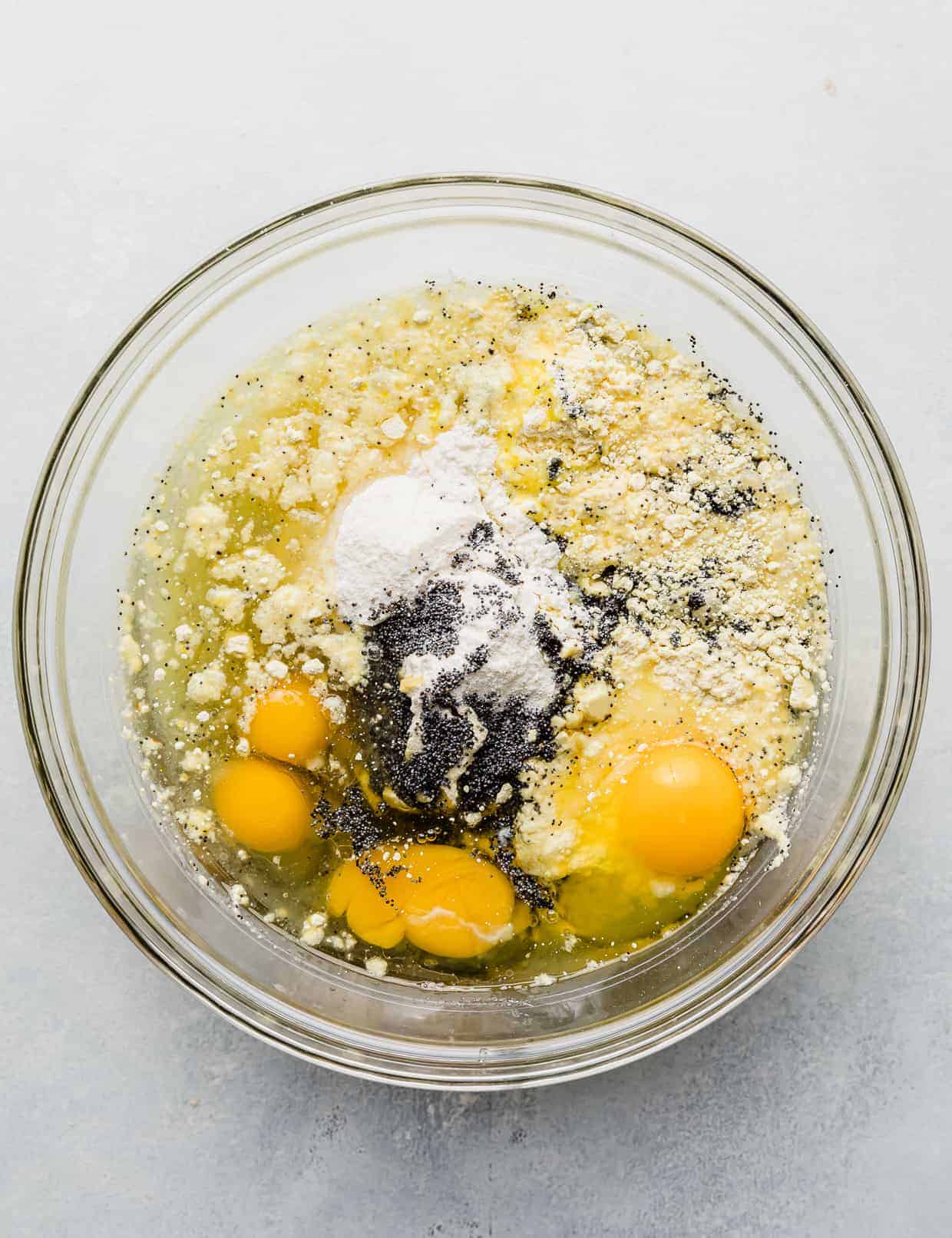 A glass bowl full of dry lemon cake mix, water, oil, 4 cracked eggs, lemon pudding mix, and poppy seeds.