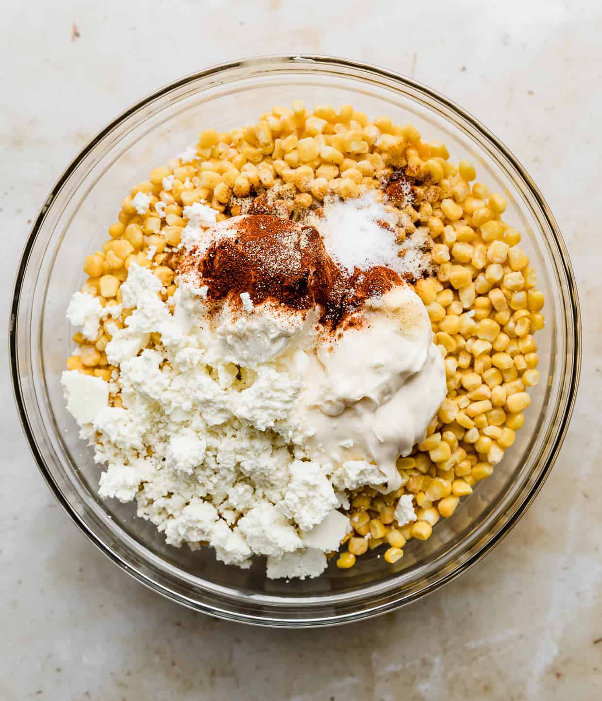 Corn kernels topped with chili powder, mayo, sour cream, and Mexican cheese in a glass bowl