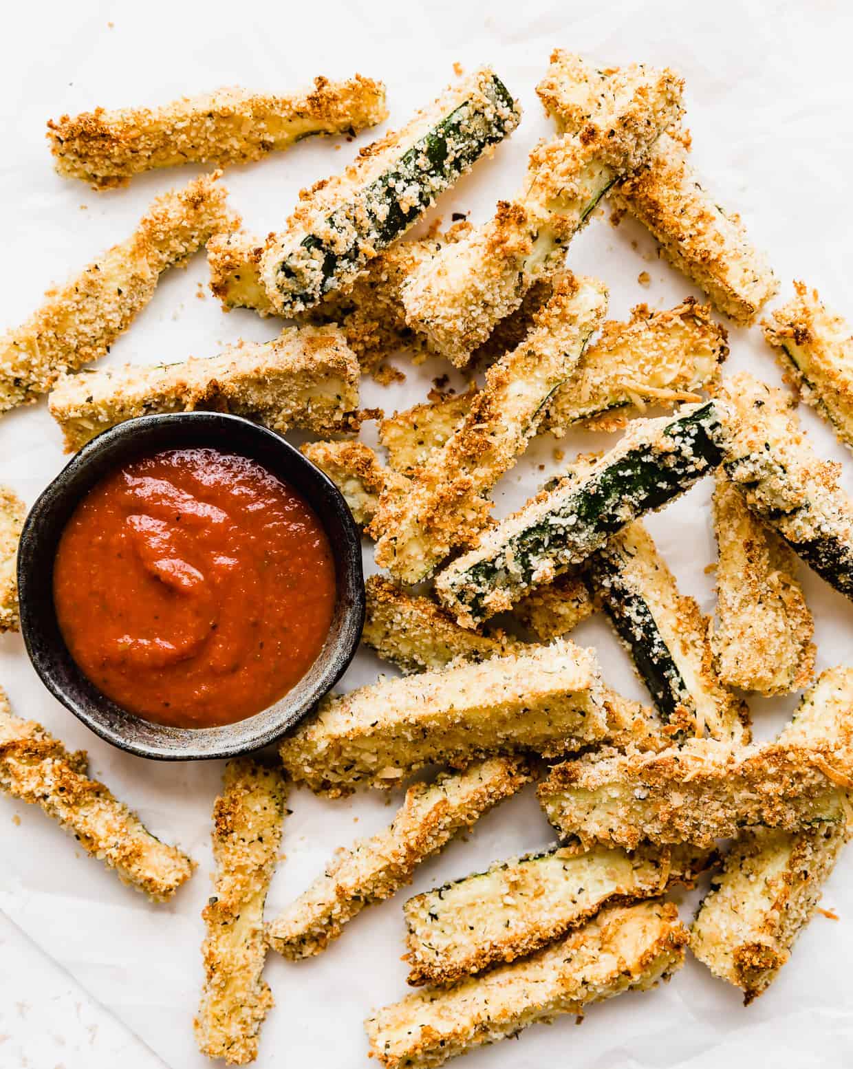 Baked Zucchini Fries on a white parchment paper with a small bowl of marinara sauce next to the fries. 