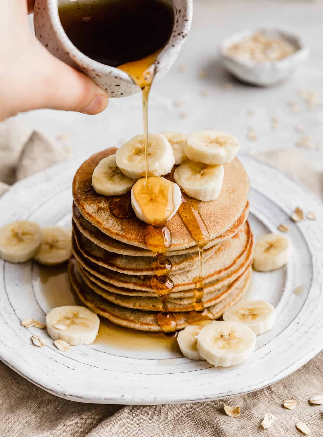 A hand pouring a white jar of maple syrup over top of Banana Oatmeal Pancakes topped with sliced bananas.