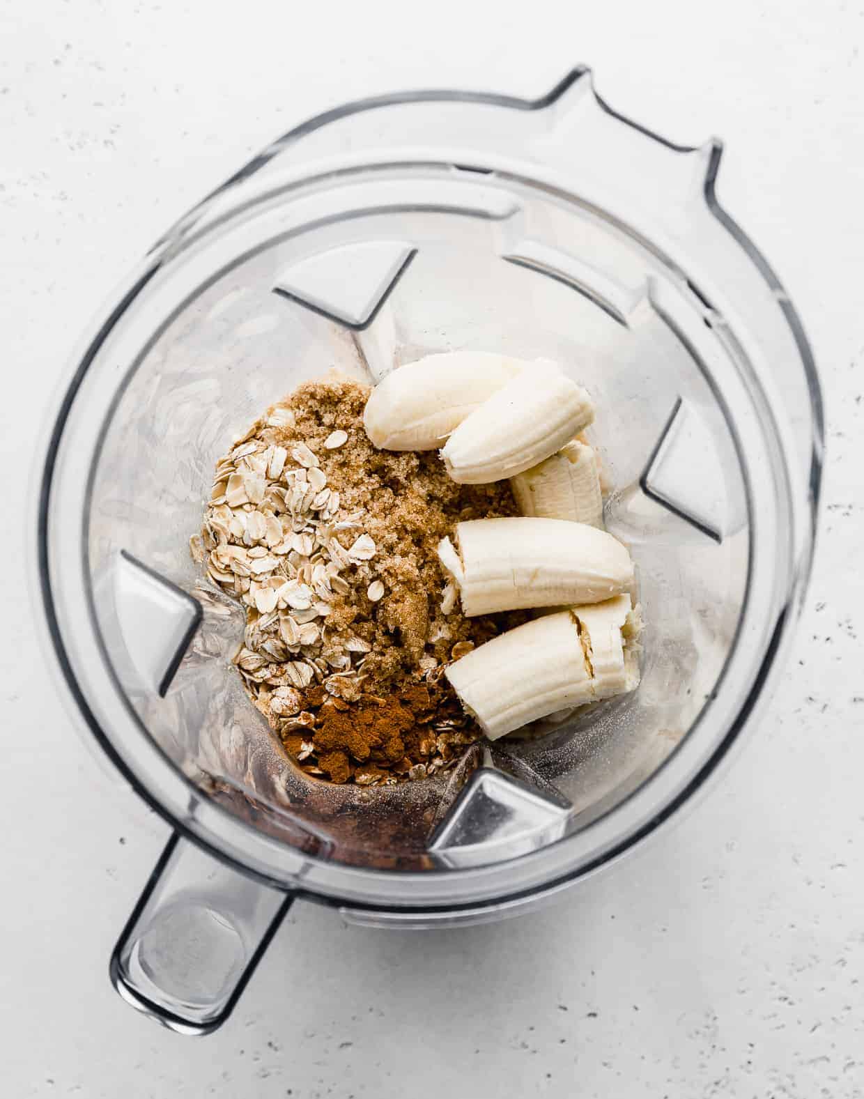 Overhead photo of a blender full of chopped bananas, oats, brown sugar, cinnamon, and milk.