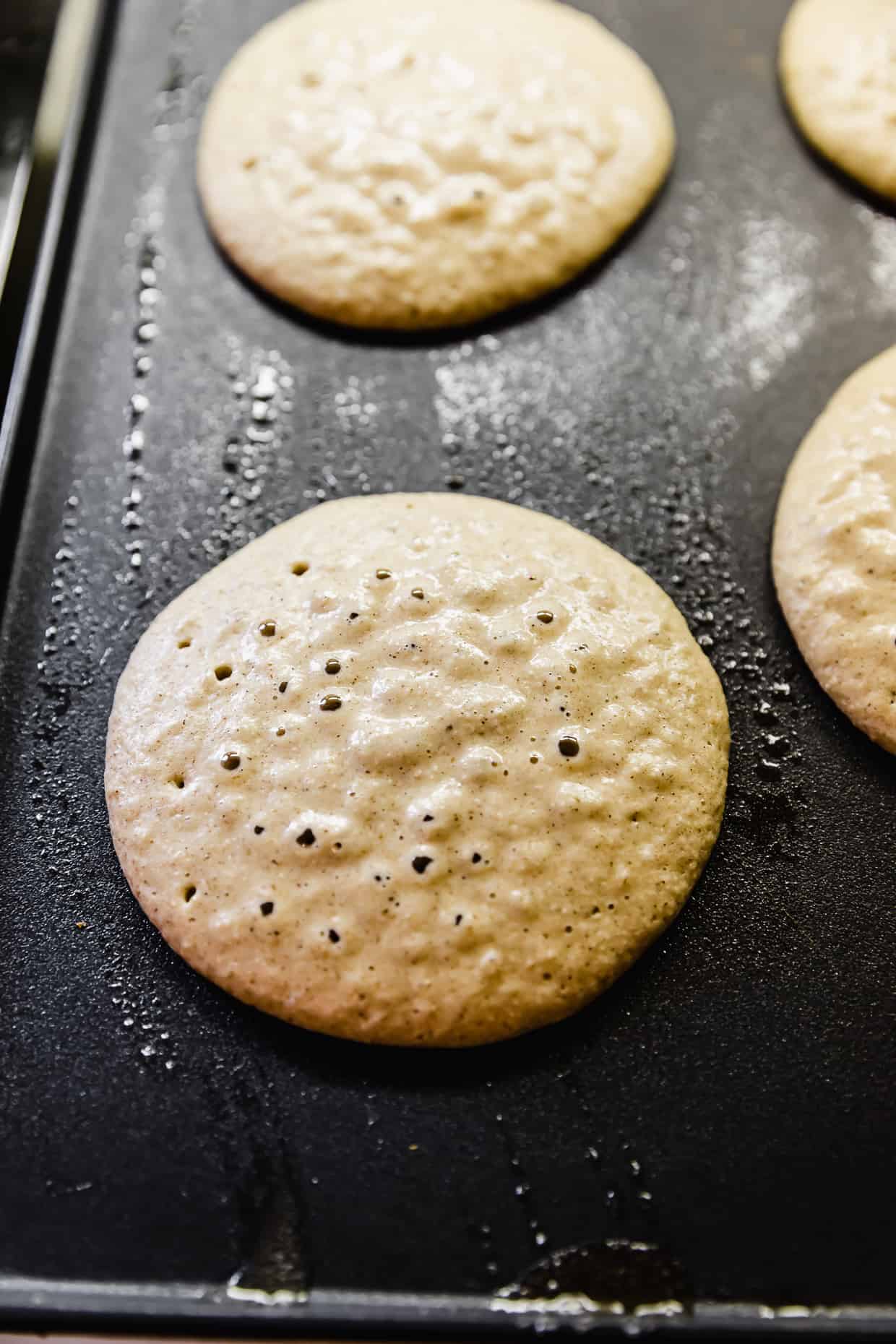 A Banana Oatmeal Pancake with bubbles oozing to the top, on a black griddle.