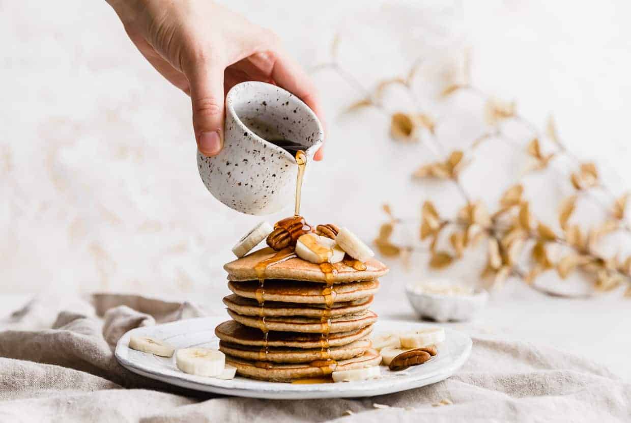 A hand pouring syrup over top a stack of Banana Oatmeal Pancakes that have sliced bananas and pecans on top. 