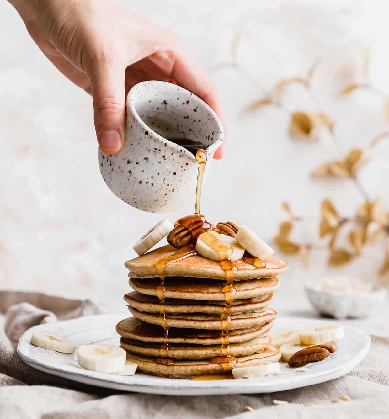 A stack of banana oatmeal pancakes on a white plate with maple syrup being drizzled overtop. 