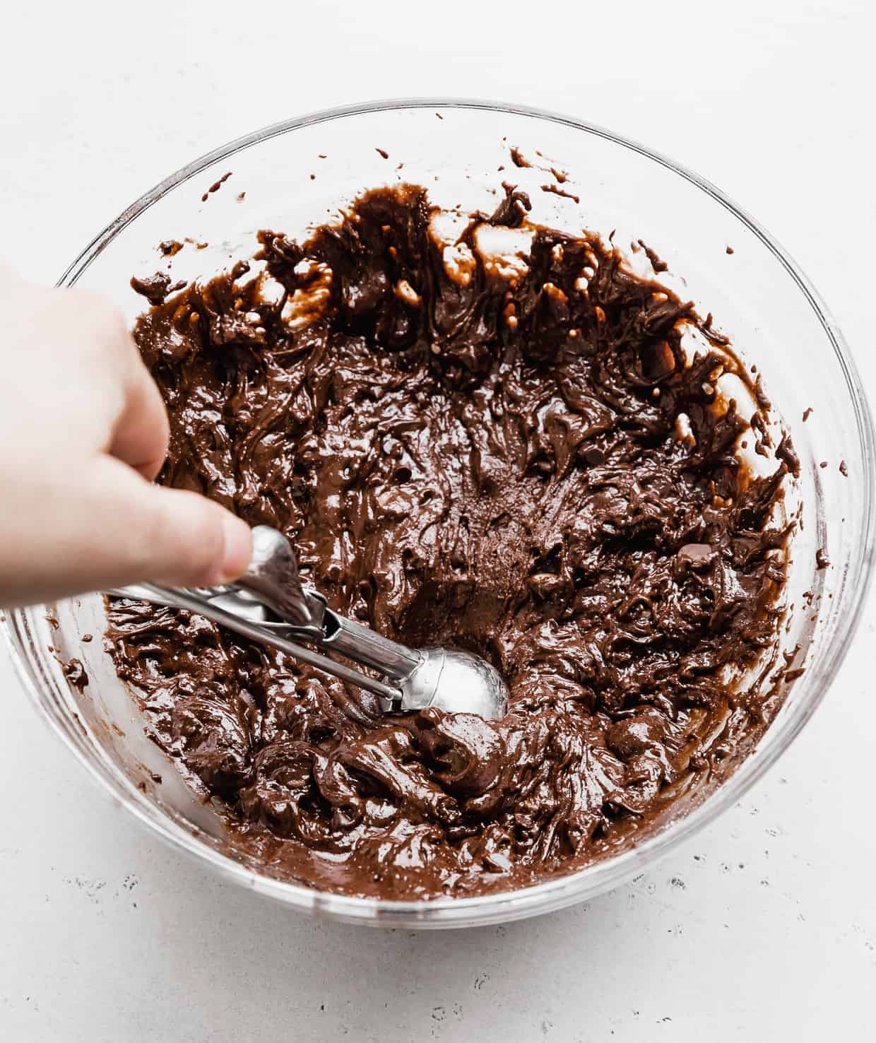 A hand holding a cookie scoop reaching into a bowl full of Brownie Mix Cookie dough batter. 