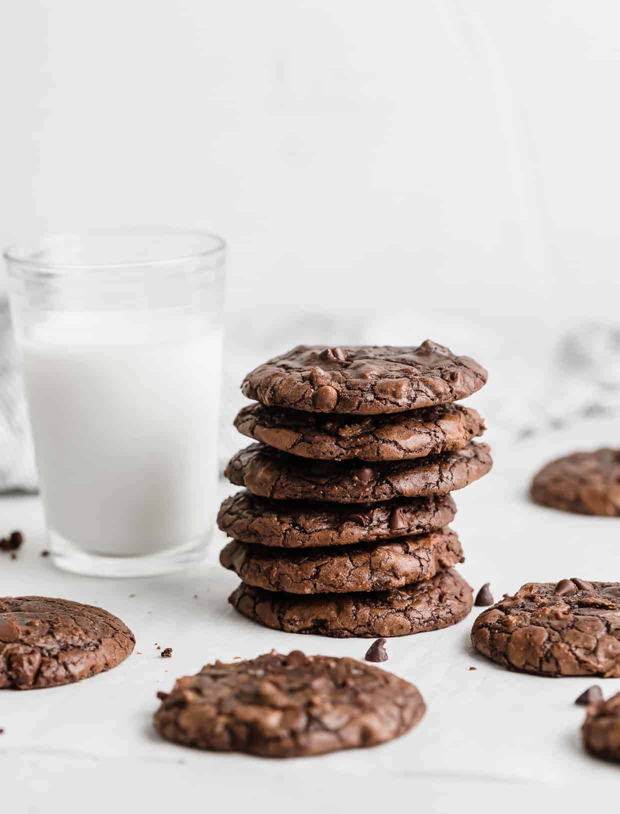 A stack of dark brown brownie cookies with a glass of milk next to it against a white background.