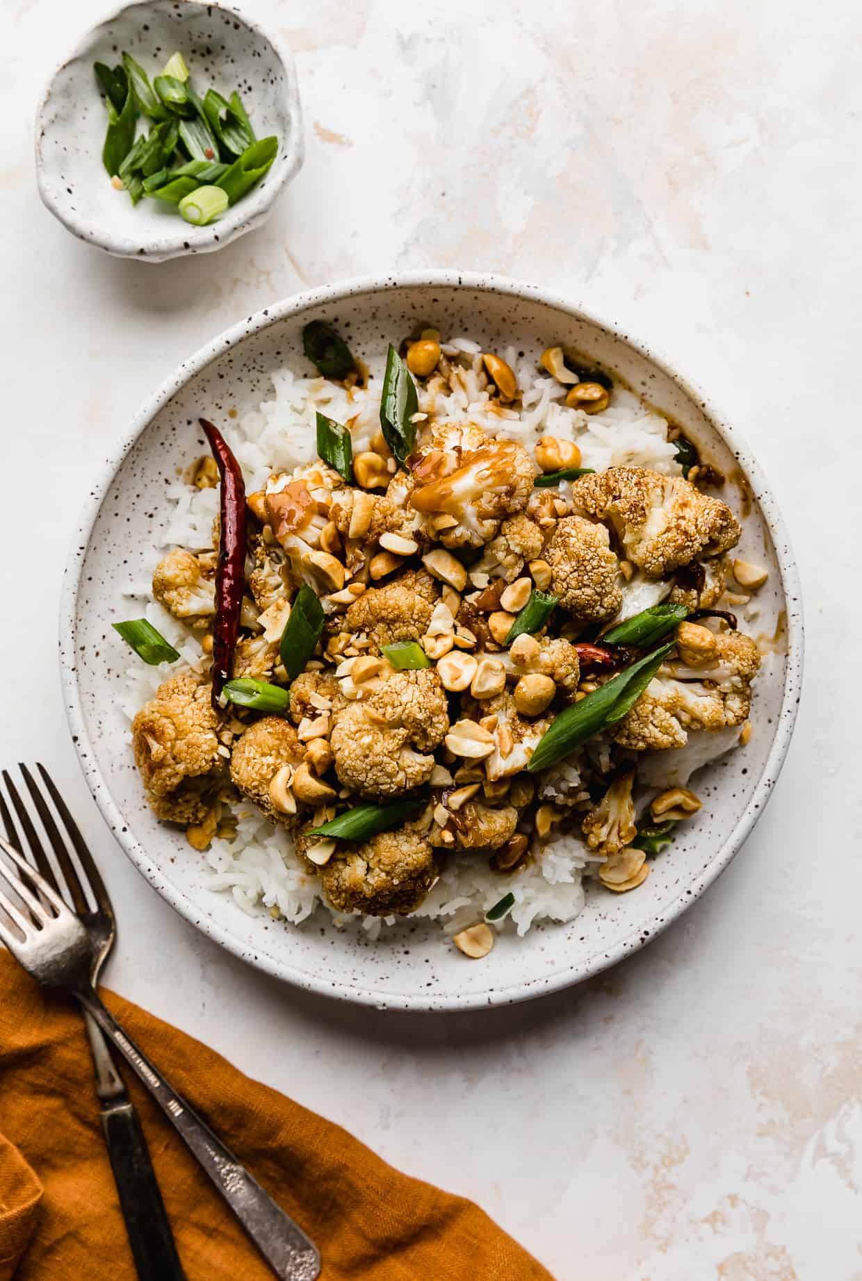 A white plate with green onion and peanut garnished Kung Pao Cauliflower, against a cream colored background.