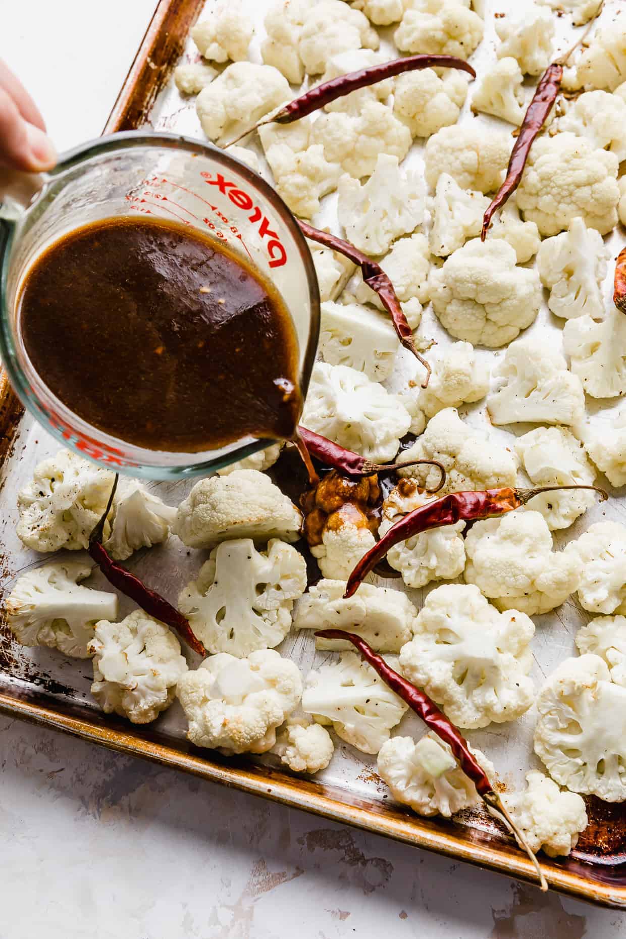 A dark red/brown liquid being poured over raw cauliflower florets on a baking sheet.