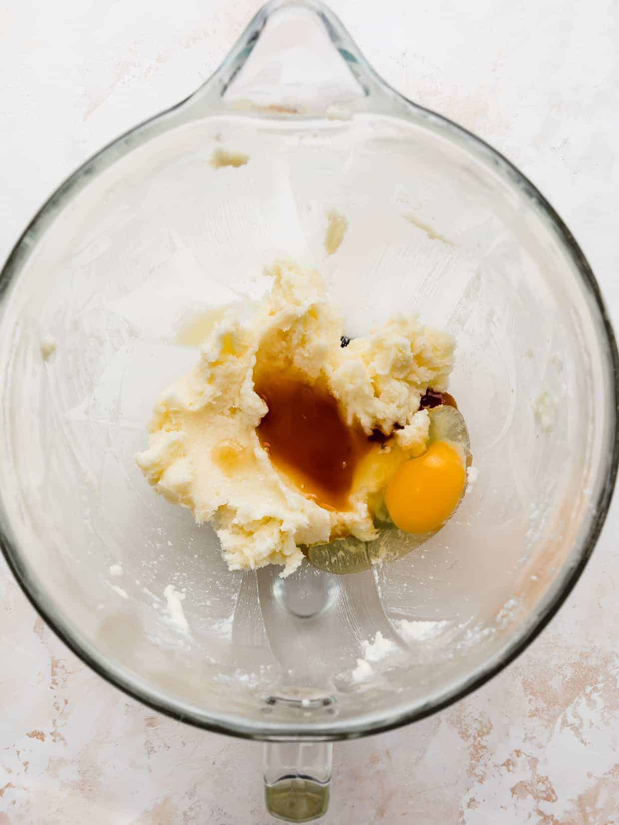 A glass bowl with creamed butter and sugar, vanilla extract, and a cracked egg in it, against a white background.