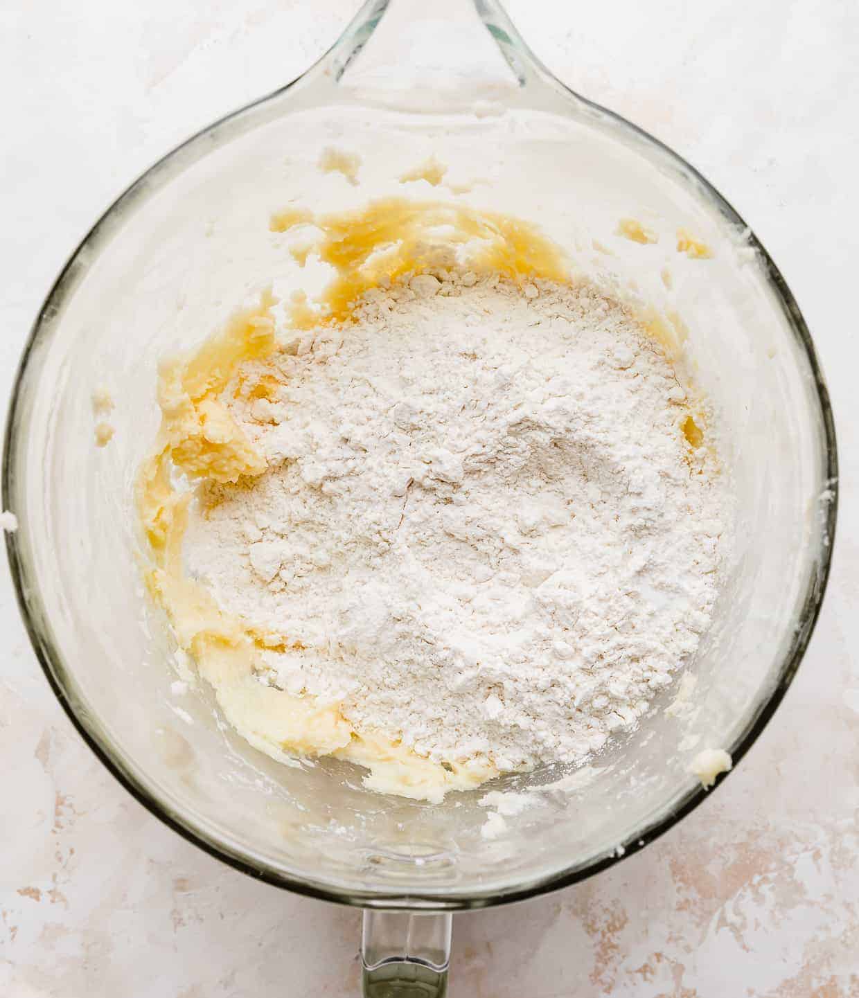A glass mixing bowl with white flour overtop of the creamed butter and sugar.