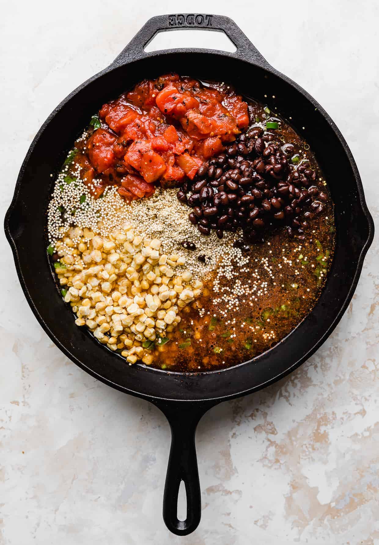 A skillet on a white background, full of uncooked quinoa, black beans, canned diced tomatoes, frozen corn kernels, and water.