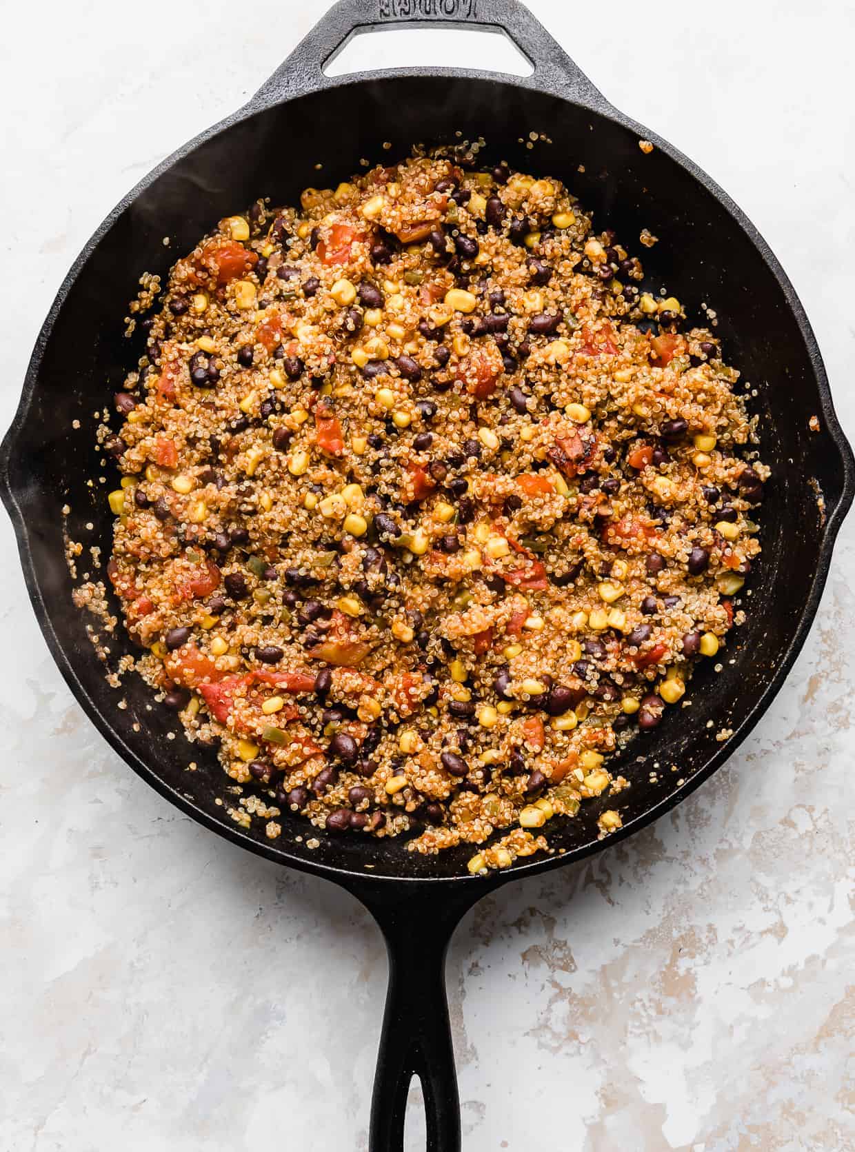 Cooked Mexican quinoa loaded with black beans, tomatoes, and corn, in a black skillet against a white background.