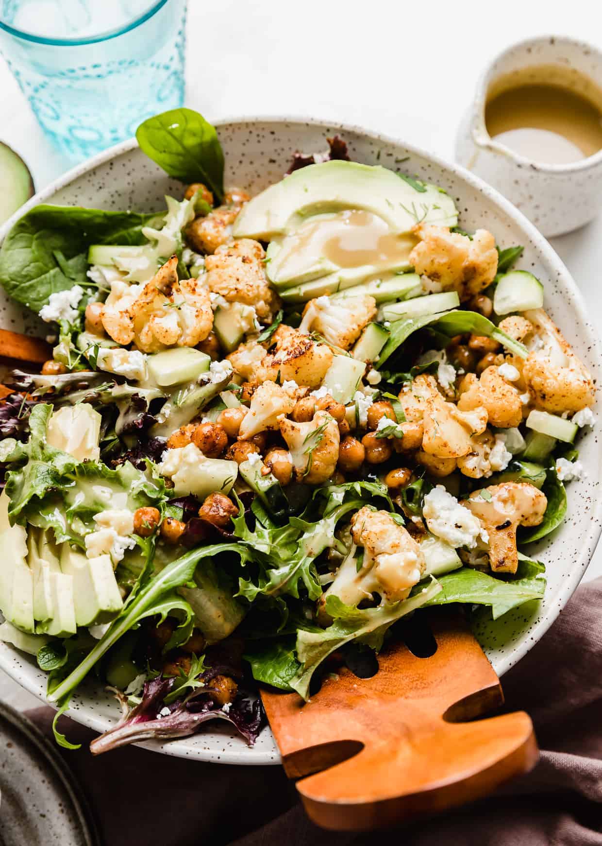 Wooden salad tongs in a large bowl that is full of mixed greens, roasted cauliflower, chickpeas, avocado, and cucumbers. 