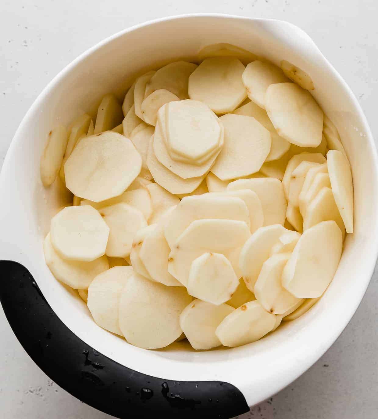 A white bowl full of raw thinly sliced potatoes.