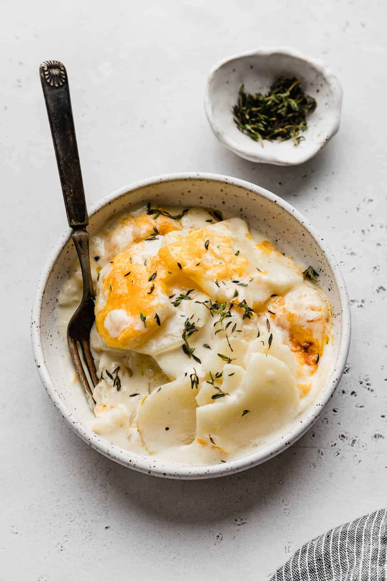 A white plate full of scalloped potatoes topped with melted cheddar cheese and fresh thyme.