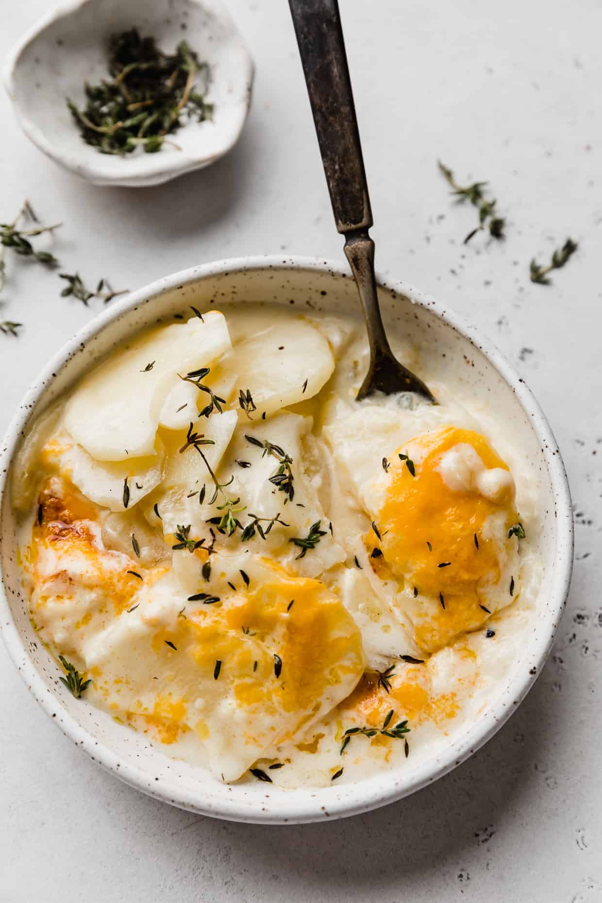 A fork amongst a plate of scalloped potatoes topped with cheddar cheese and fresh thyme, against a light gray background.