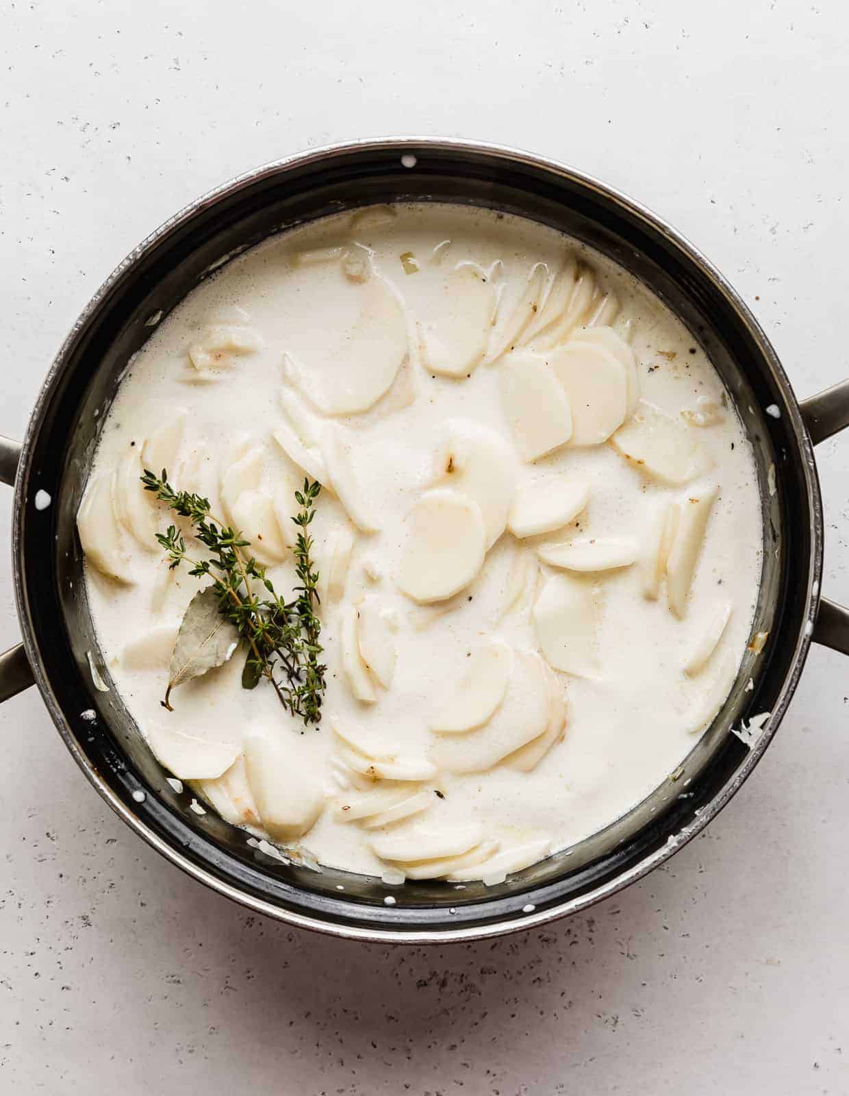 A large black pot full of heavy cream, thinly sliced potatoes, sprigs of thyme, and bay leaves.