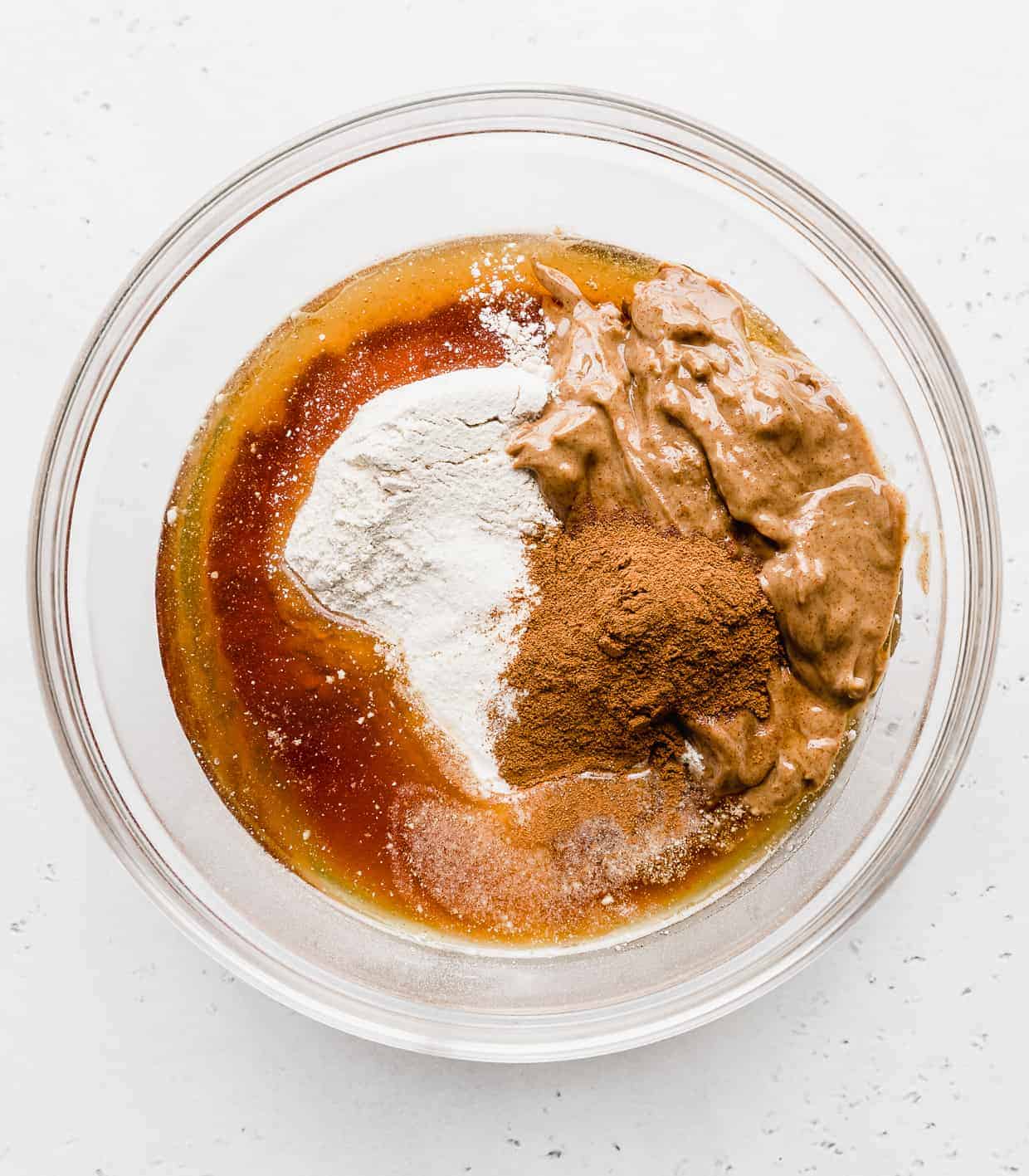 A glass bowl full of ingredients used to make Snickerdoodle Energy Bites; almond butter, maple syrup, protein powder, cinnamon, and kodiak cakes mix. 
