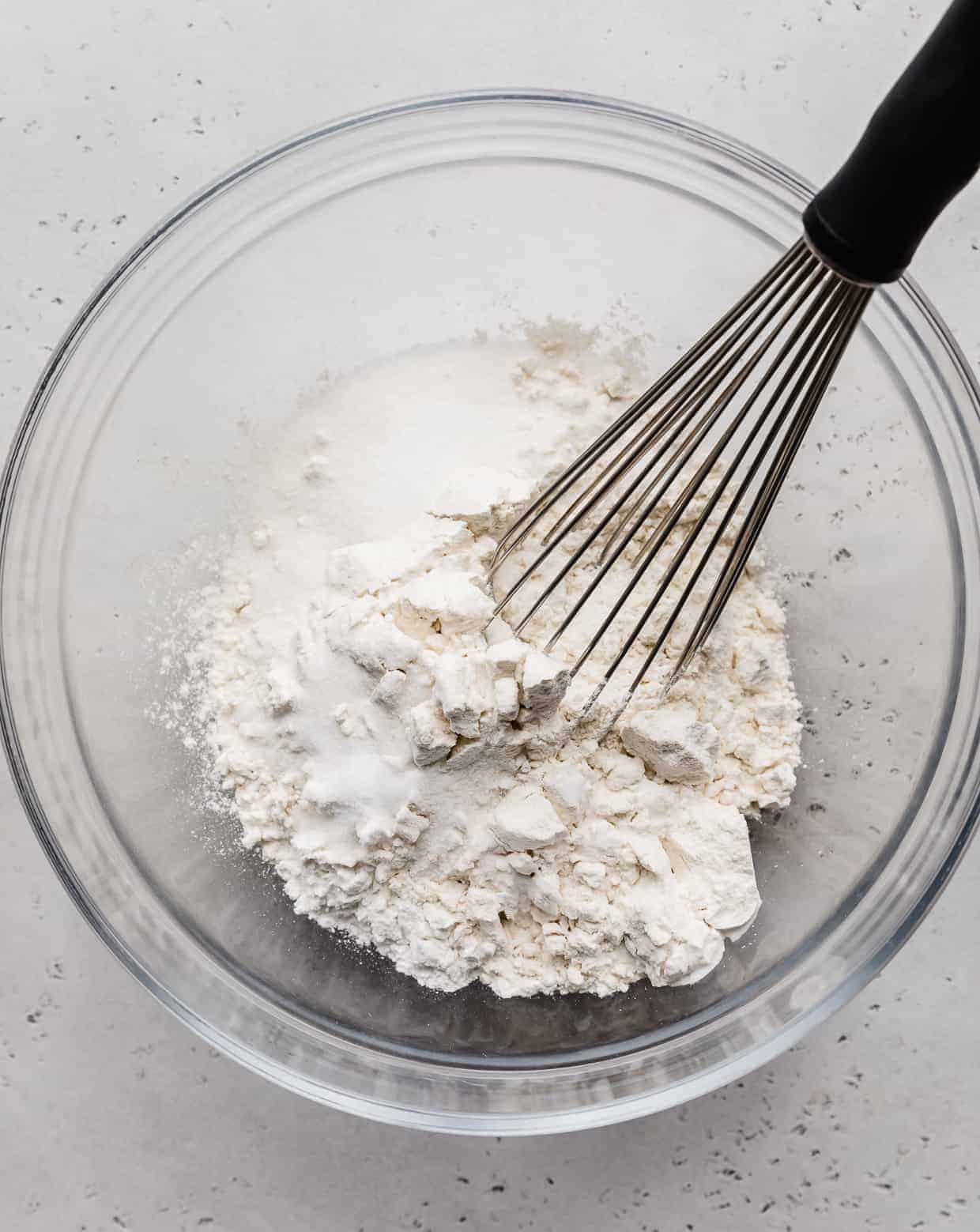 A glass bowl with a whisk and flour in it.