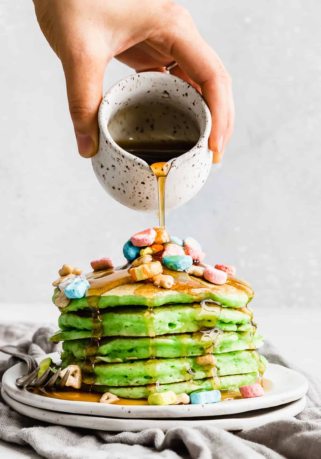 A stack of green pancakes on a plate topped with lucky charms cereal with maple syrup being poured overtop. 