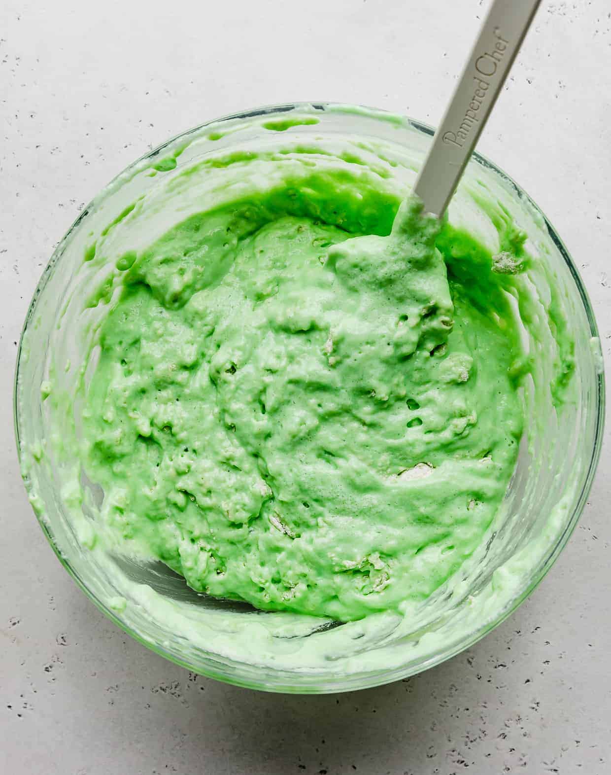 A thick green pancake batter in a glass bowl against a light gray background. 