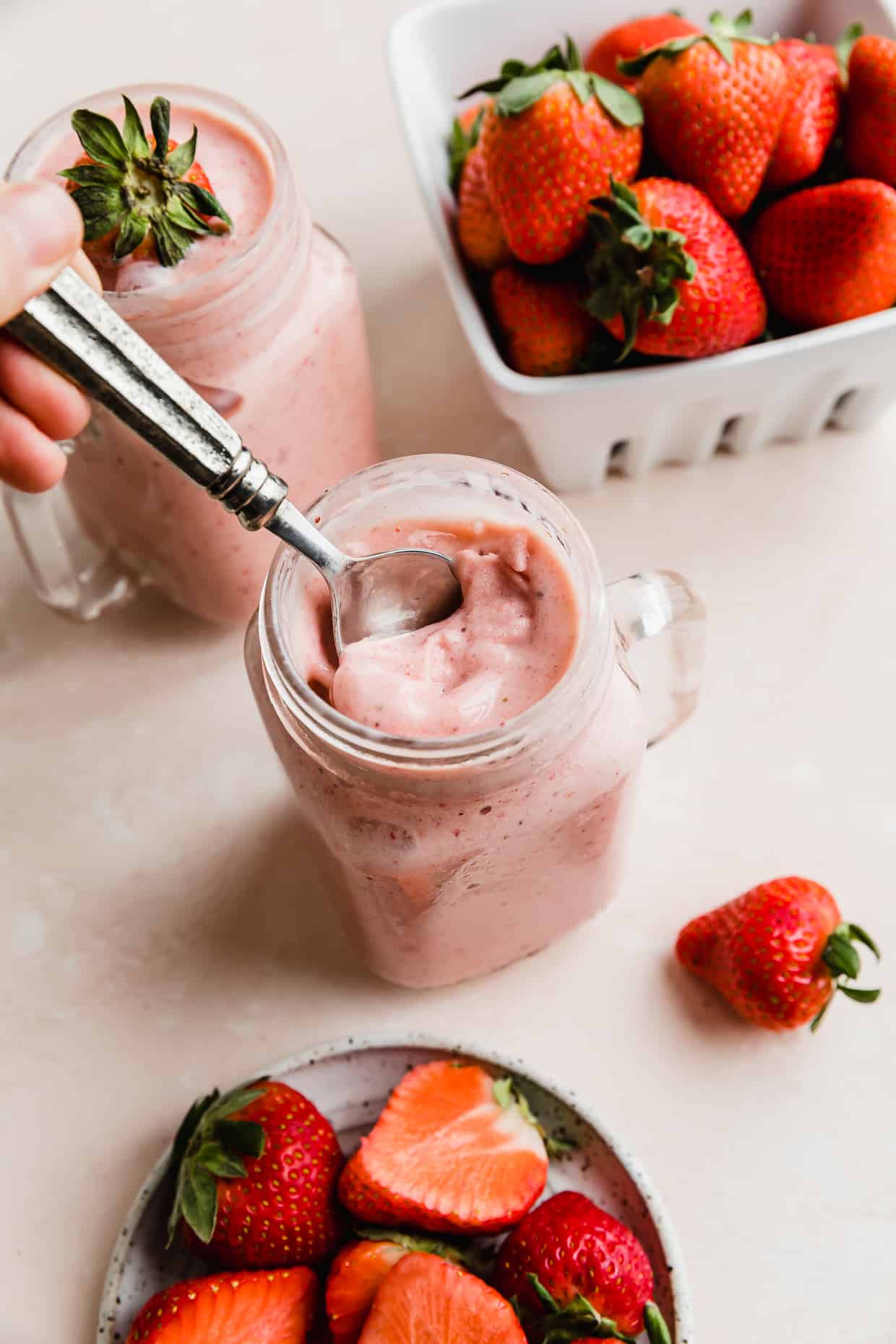 A spoon scooping up a portion of a strawberry pineapple smoothie that's in a mason glass jar, with a basket of strawberries in the background.