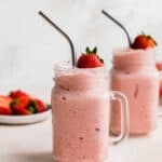 Two mason jar glasses full of Strawberry Pineapple Smoothie with a fresh strawberry resting on the top of the smoothie.