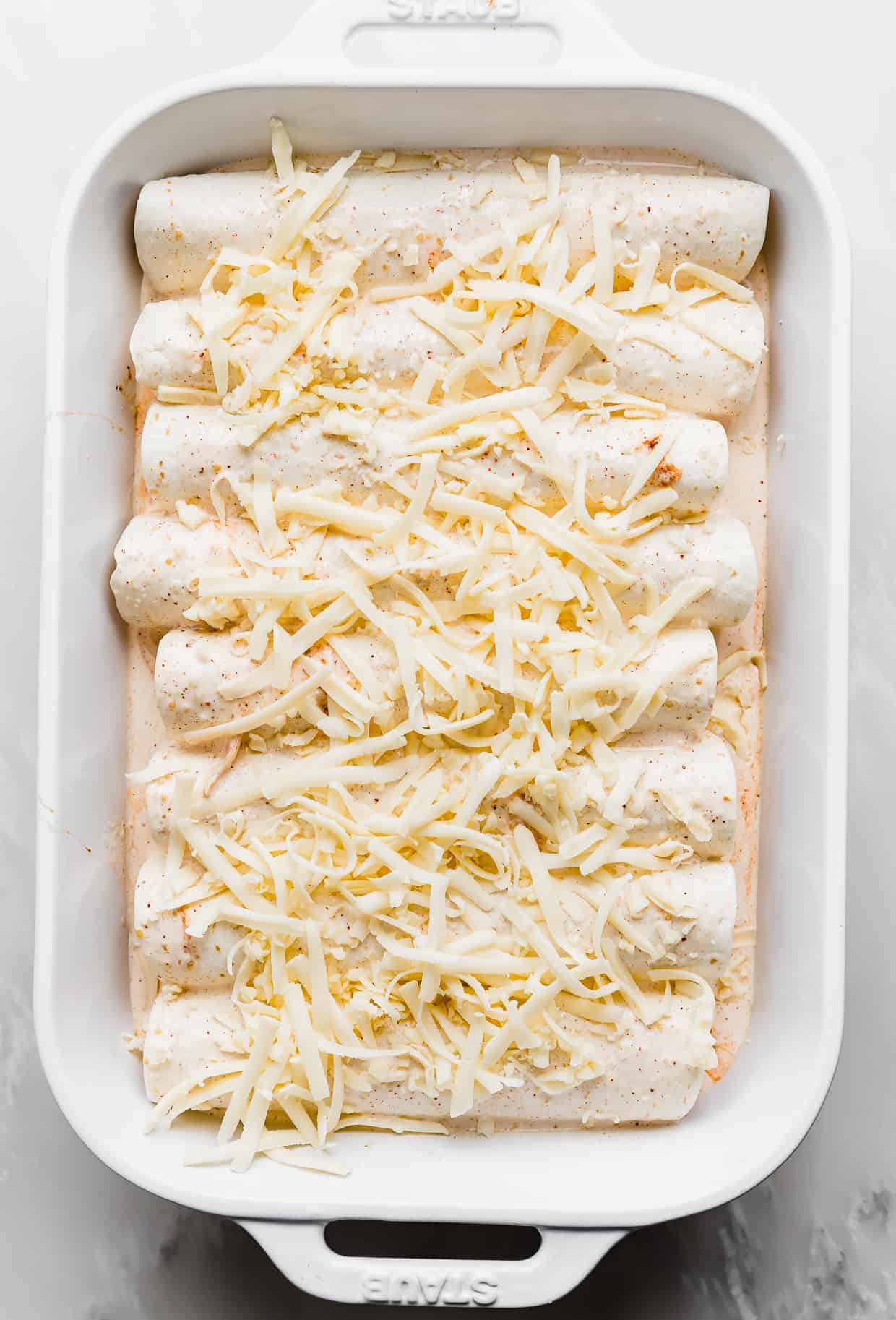 White shredded cheese overtop rolled up flour tortilla enchiladas in a casserole dish.