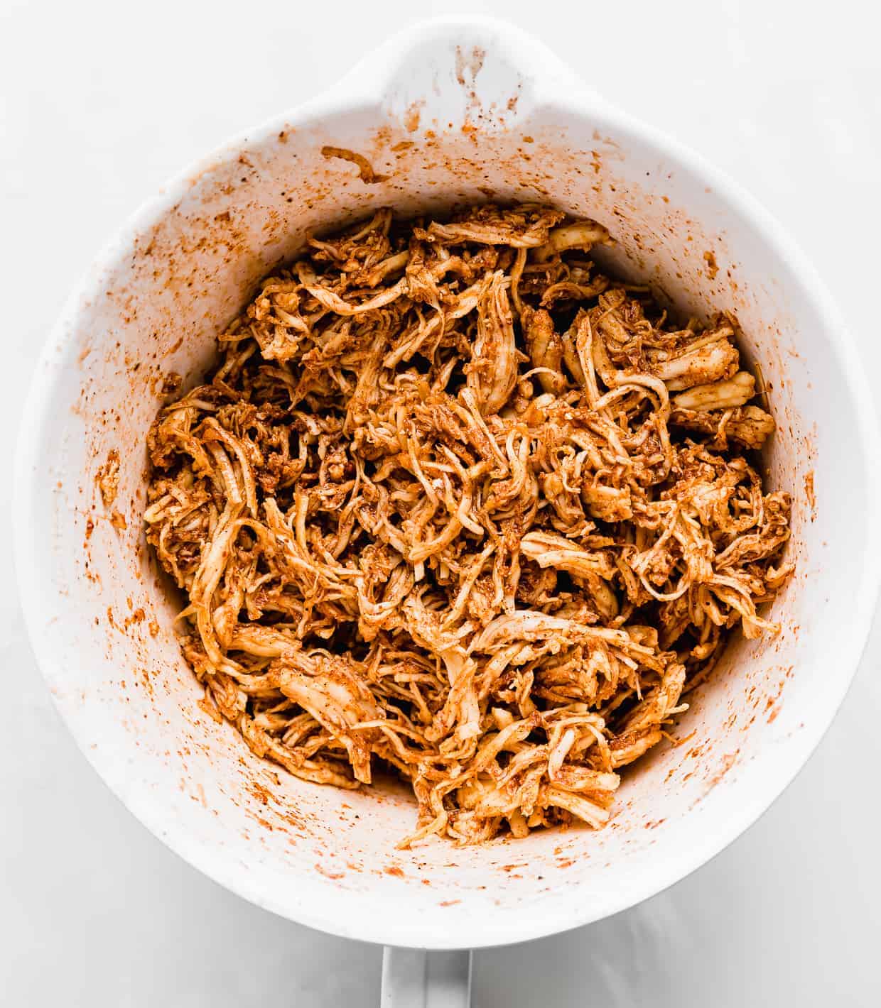 A white bowl full of shredded chicken covered in a red and orange colored marinade. 