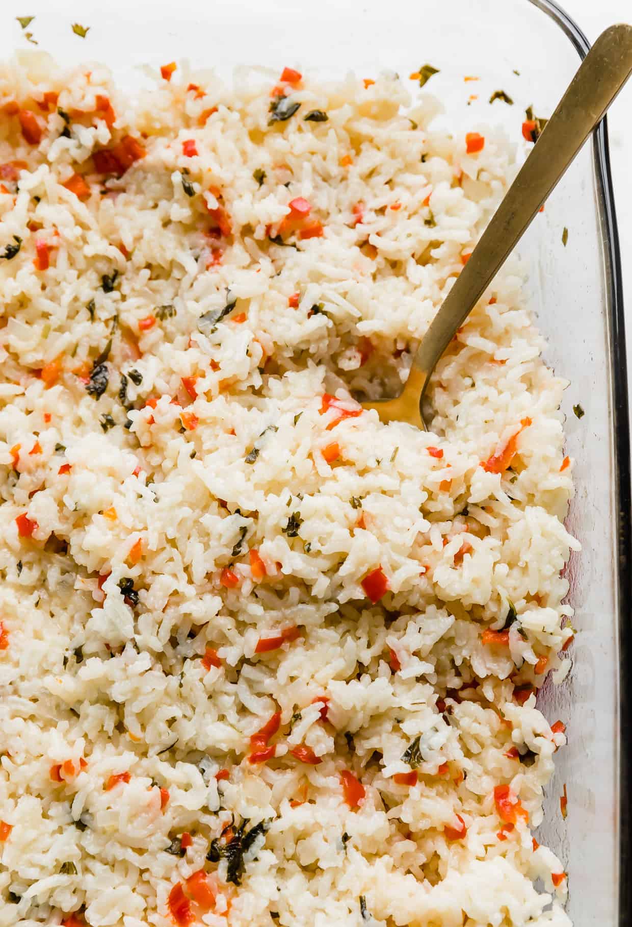 A spoon in a baking dish full of Sweet Pepper Rice that is studded with diced red peppers and chopped cilantro. 