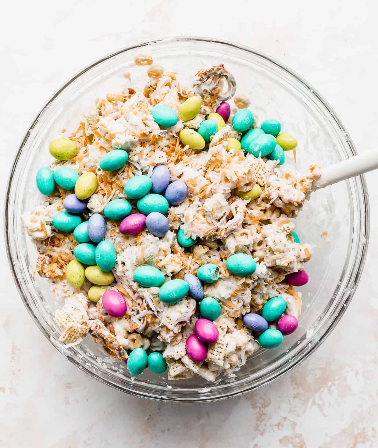 Easter (pastel) colored M&M's being stirred into a cereal trail mix in a glass bowl. 