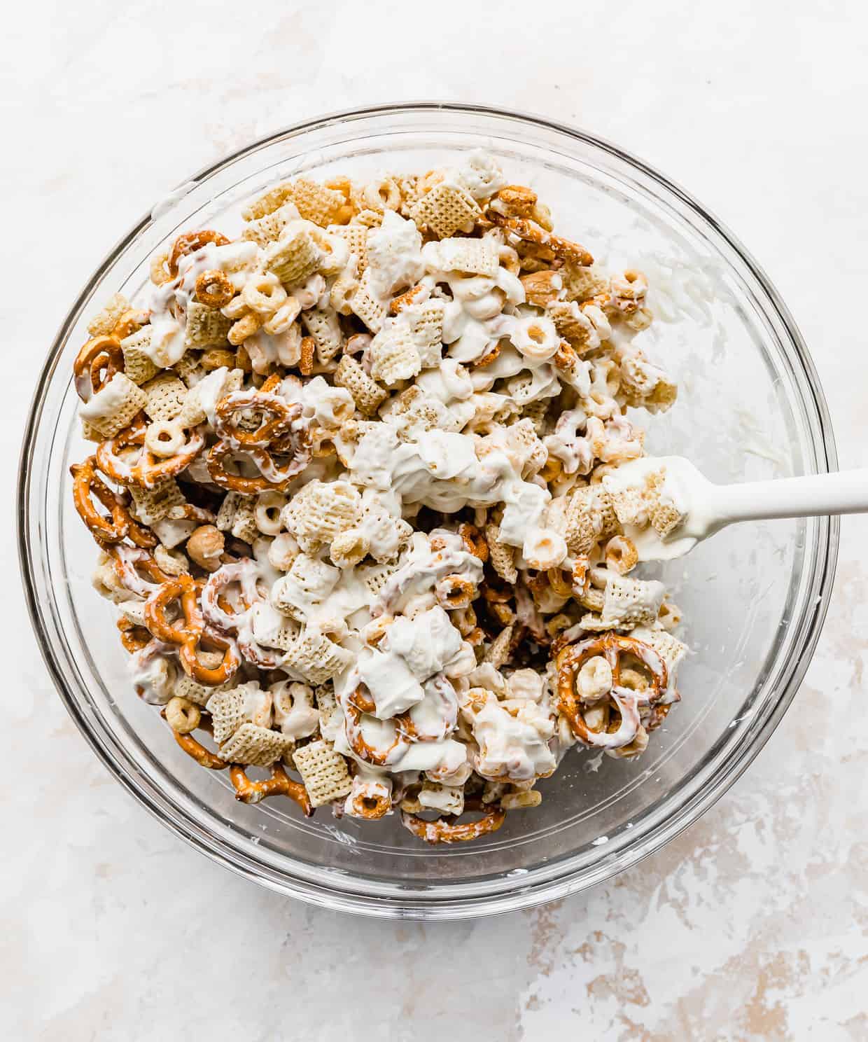 White chocolate being stirred into a glass bowl with pretzels, cashews, cheerios.