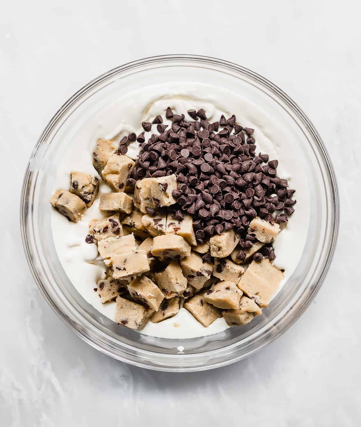 Vanilla ice cream, cookie dough bites, and mini chocolate chips in a glass bowl against a white background.