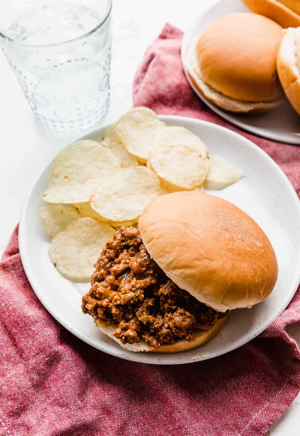 A homemade sloppy Joe on a white plate with potato chips next to the sandwich. 